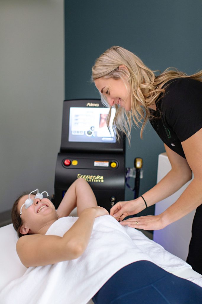 A woman lies on a treatment table with protective eyewear, smiling as she receives an aesthetics laser procedure on her underarm from another smiling woman. A laser device with a screen is in the background.