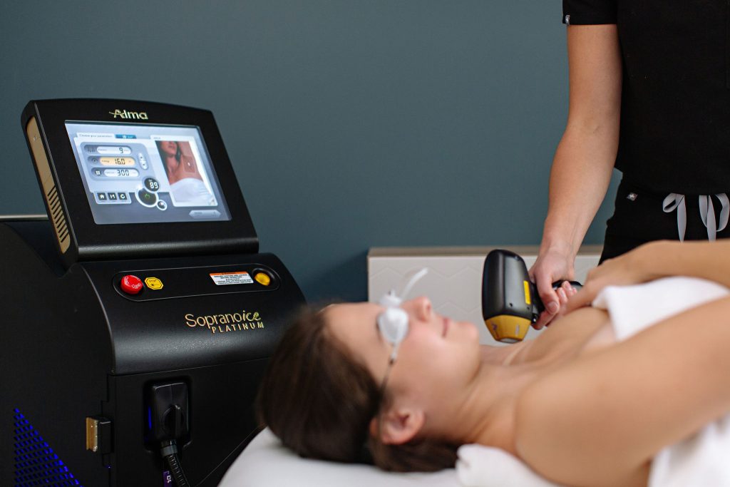 A person lies on a treatment table with protective eyewear, receiving a laser hair removal treatment for enhanced aesthetics on their underarm area. The technician holds a handheld laser device connected to a machine with a display screen showing treatment settings.