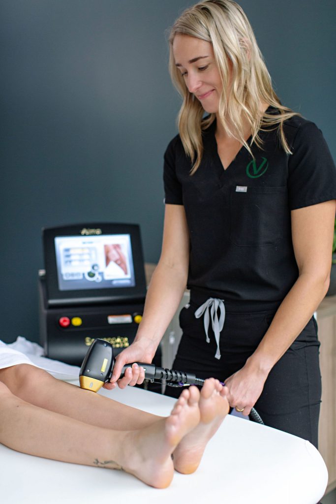 A person with long, blonde hair wearing a black scrub top and gray drawstring pants operates a laser hair removal device on a client's legs. The client is lying on a treatment table, enhancing their aesthetics, with a monitor displaying treatment settings in the background.