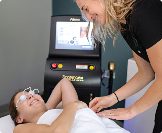 A person lies on a treatment table with a towel covering them while wearing protective eyewear. They are smiling as a technician in a black shirt, standing beside them, operates a laser machine in the background. The setting exudes aesthetics, enhancing the ambiance of this cosmetic treatment room.